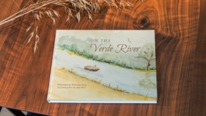 Book on table. "On the Verde River" by Phoebe Fox and illustrated in watercolor by Jim Fox Cover is boy rowing down river in boat with lush green and sand on the banks