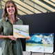 Author Phoebe Fox of Mamafoxbooks holds a copy of the book On the Verde River, standing in front of Jim Fox watercolor